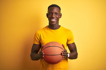 African american athlete man holding basketball ball standing over isolated yellow background with a happy face standing and smiling with a confident smile showing teeth