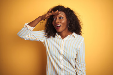 African american woman wearing striped shirt standing over isolated yellow background very happy and smiling looking far away with hand over head. Searching concept.