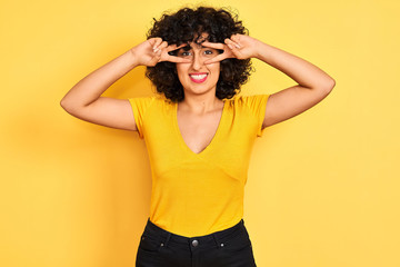 Young arab woman with curly hair wearing t-shirt standing over isolated yellow background Doing peace symbol with fingers over face, smiling cheerful showing victory