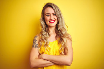 Young beautiful woman wearing t-shirt standing over yellow isolated background happy face smiling with crossed arms looking at the camera. Positive person.