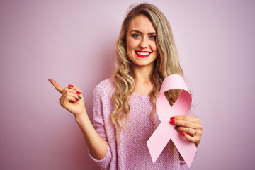 Young beautiful woman holding cancer ribbon over pink isolated background very happy pointing with...