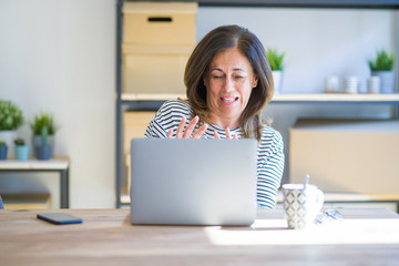 Middle age senior woman sitting at the table at home working using computer laptop disgusted expression, displeased and fearful doing disgust face because aversion reaction. With hands raised. 