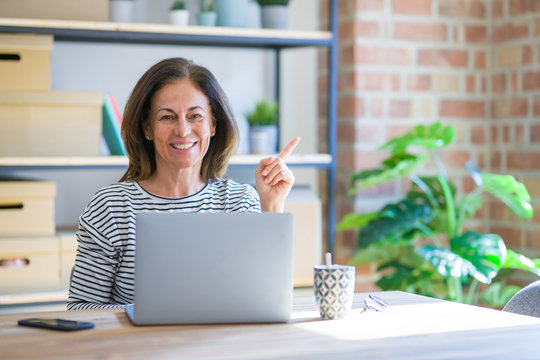 Middle age senior woman sitting at the table at home working using computer laptop very happy pointing with hand and finger to the side