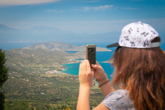 Young woman making photo of a beautiful background of aegean sea and mountains. Woman with brown wavy hair taking picture on vacations.