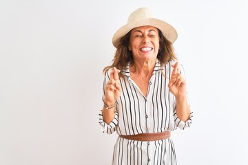 Obraz na płótnie Canvas Middle age businesswoman wearing striped dress and hat over isolated white background gesturing finger crossed smiling with hope and eyes closed. Luck and superstitious concept.