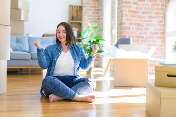 Young plus size woman sitting on the floor around cardboard boxes moving to a new home celebrating surprised and amazed for success with arms raised and open eyes. Winner concept.