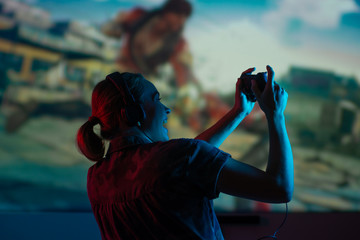 A girl gamer plays games with a gamepad, joystick on a large screen, with bright light and a dark room. eSports, gaming, streaming.bright background