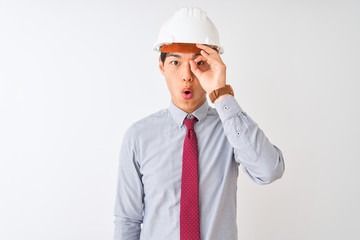 Chinese architect man wearing tie and helmet standing over isolated white background doing ok gesture shocked with surprised face, eye looking through fingers. Unbelieving expression.