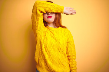 Beautiful redhead woman wearing winter sweater standing over isolated yellow background covering eyes with arm, looking serious and sad. Sightless, hiding and rejection concept