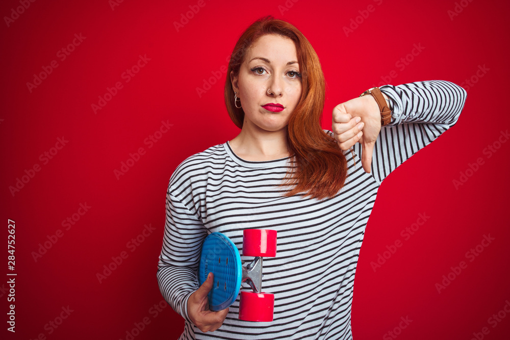 Wall mural young redhead student woman holding skate over red isolated background with angry face, negative sig