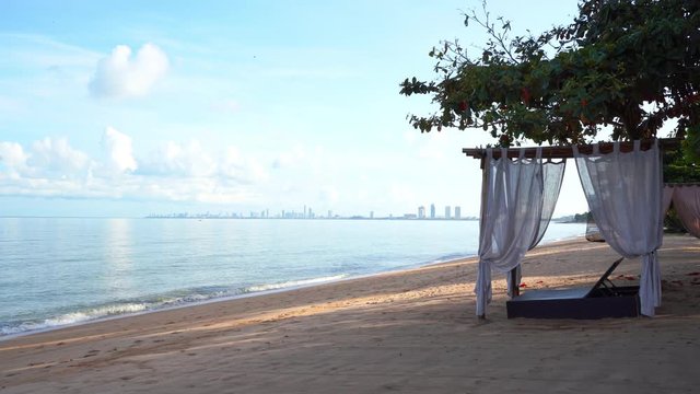 An empty cabana sits on an empty beach facing the lagoon water and looking off to the modern city skyline in the distance.