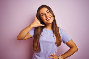Fototapeta premium Young beautiful woman wearing casual t-shirt standing over isolated pink background smiling doing phone gesture with hand and fingers like talking on the telephone. Communicating concepts.
