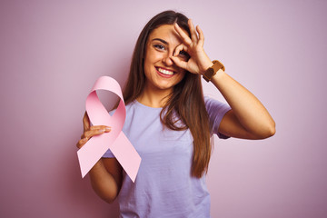 Young beautiful woman holding cancer ribbon standing over isolated pink background with happy face...