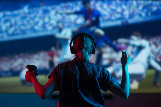 Soccer game, girl gamer playing a game in football headphones on a big screen, with bright light and a dark room. Gameplay, streaming, e-sports.