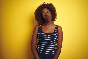 Young african afro woman wearing striped t-shirt over isolated yellow background making fish face with lips, crazy and comical gesture. Funny expression.