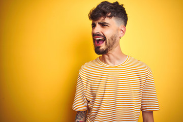 Young man with tattoo wearing striped t-shirt standing over isolated yellow background angry and mad screaming frustrated and furious, shouting with anger. Rage and aggressive concept.