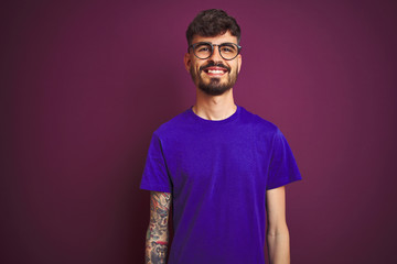 Young man with tattoo wearing t-shirt and glasses standing over isolated purple background with a happy and cool smile on face. Lucky person.