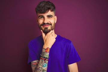 Young man with tattoo wearing t-shirt standing over isolated purple background looking confident at the camera smiling with crossed arms and hand raised on chin. Thinking positive.