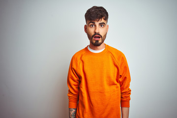 Young man with tattoo wearing orange sweater standing over isolated white background afraid and shocked with surprise expression, fear and excited face.