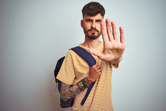 Young student man with tattoo wearing backpack standing over isolated white background with open hand doing stop sign with serious and confident expression, defense gesture