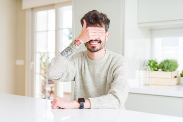 Young man wearing casual sweater sitting on white table smiling and laughing with hand on face covering eyes for surprise. Blind concept.