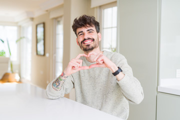 Fototapeta premium Young man wearing casual sweater sitting on white table smiling in love showing heart symbol and shape with hands. Romantic concept.