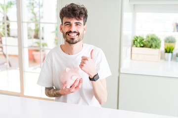 Fototapeta na wymiar Young man holding piggy bank as insurance investment happy with big smile doing ok sign, thumb up with fingers, excellent sign