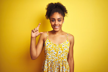 African american woman wearing casual floral dress standing over isolated yellow background smiling and confident gesturing with hand doing small size sign with fingers looking and the camera. 