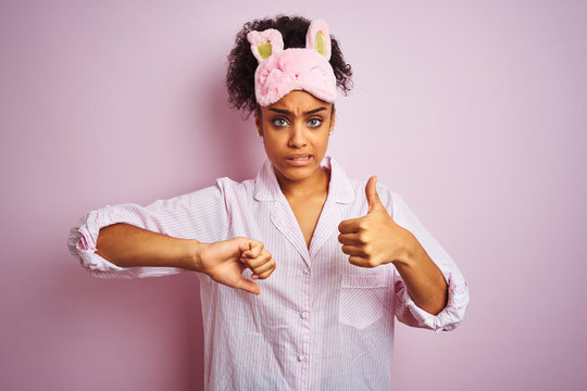 Young african american woman wearing pajama and mask over isolated pink background Doing thumbs up and down, disagreement and agreement expression. Crazy conflict