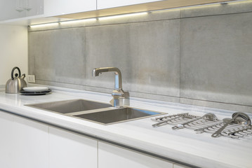 Detail of a white rectangular designer kitchen sink with chrome water tap against a tiled wall