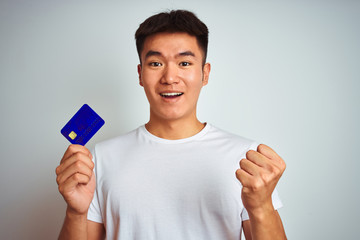 Young asian chinese man holding credit card standing over isolated white background screaming proud and celebrating victory and success very excited, cheering emotion