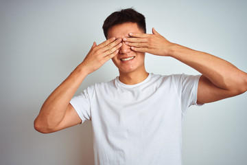 Young asian chinese man wearing t-shirt standing over isolated white background covering eyes with hands smiling cheerful and funny. Blind concept.