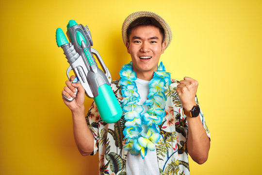 Asian chinese man wearing floral hawaian lei and water gun over isolated yellow background screaming proud and celebrating victory and success very excited, cheering emotion