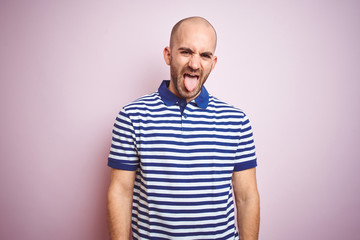 Obraz premium Young bald man with beard wearing casual striped blue t-shirt over pink isolated background sticking tongue out happy with funny expression. Emotion concept.