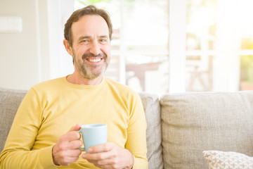 Handsome middle age man sitting on the sofa relaxed drinking a cup of coffe and smiling at the camera at home