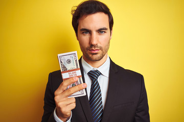 Young handsome businessman wearing suit holding dollars over isolated yellow background with a confident expression on smart face thinking serious