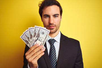 Young handsome businessman wearing suit holding dollars over isolated yellow background with a confident expression on smart face thinking serious