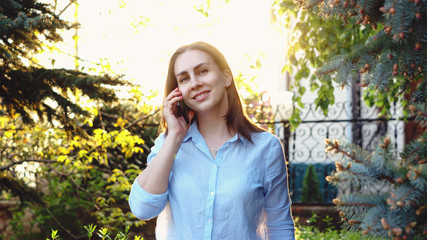 Beautiful woman talking by phone with friend while standing in the park, smiling hipster girl using...