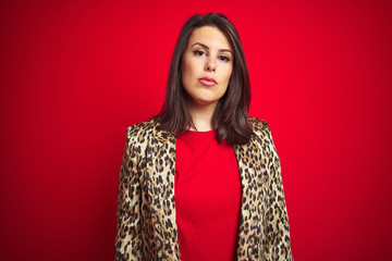 Young beautiful brunette woman wearing a leopard jacket over red isolated background with serious...