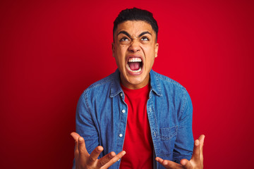 Young brazilian man wearing denim shirt standing over isolated red background crazy and mad...