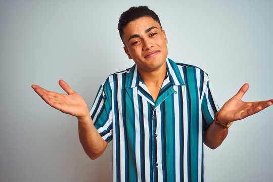 Young brazilian man wearing summer striped shirt standing over isolated white background clueless and confused expression with arms and hands raised. Doubt concept.