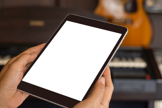 Mockup image of hands holding and using black tablet computer with blank white screen in music room with guitar and piano blurred background. clipping path - Powered by Adobe
