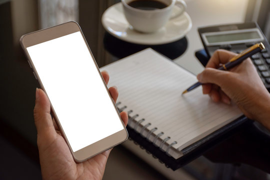 Mockup image of hand holding mobile smartphone with blank white screen and writing on empty white note paper with calculator and coffee cup on the desk. Workspace for web design montage. 