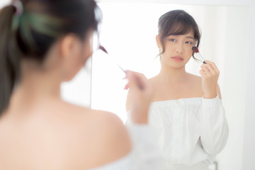 Beauty portrait young asian woman smiling with face looking mirror applying makeup with brush cheek in the bedroom, beautiful girl holding blusher, skin care and cosmetic table fashion concept.