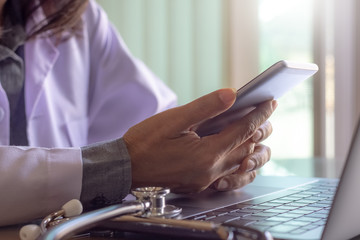 Female doctor in white lab coat using mobile smartphone in free time with laptop computer with medical stethoscope on the desk in medical room at clinic or hospital.	