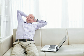 Smiling lazy middle-aged man sitting on his sofa and stretching back instead of working on laptop