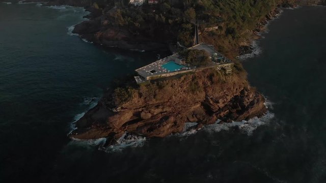 Top down aerial pan showing a recreational construction on a small island on the coast of Rio de Janeiro seen from above at golden hour sunrise with umbrellas casting a shade around a pool
