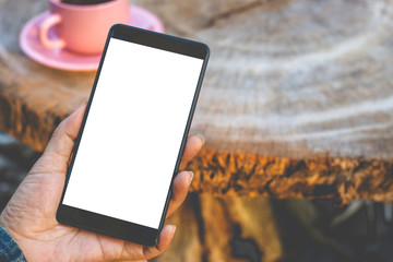 Mockup woman hand holding black mobile smart phone with blank white screen with pink cup of coffee on wooden table.