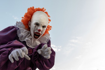 Scary clown against the sky. A teenage boy in a suit like a red-haired Pennywise opened his mouth with sharp teeth close-up. Cosplay IT for carnival or for halloween. Copy space.