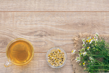 Glass cup of green tea with dry and fresh white chamomile flowers on wooden background.